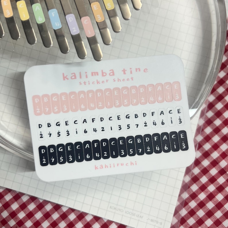 Pu&ograve; includere: Un foglio di adesivi per lamelle di kalimba con tre file di etichette con note musicali e numeri. Il foglio &egrave; bianco con dettagli rosa e neri. Il testo "kalimba tine sticker sheet" &egrave; visibile.