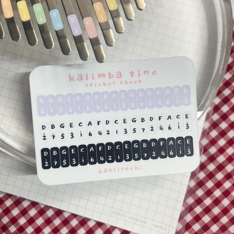 Pu&ograve; includere: Un foglio di adesivi per kalimba con note musicali e il testo "kalimba tine sticker sheet" in rosa. Il foglio di adesivi ha tre file di note, con il nome del marchio "kahiiruchi" in basso. Le lamelle del kalimba sono visibili sullo sfondo.