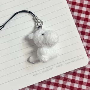 May include: A white plush dog charm with a black string and silver-tone metal clasp. The small dog is sitting on a lined paper notebook. The notebook has the text "KOKUYO LOOSE-LEAF -807BT 6 mm ruled x 28 lines".