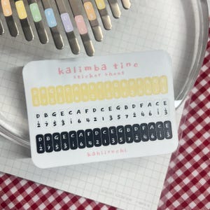 Pu&ograve; includere: Un foglio di adesivi bianco con l'etichetta "kalimba tine" con note musicali e numeri. Il foglio di adesivi ha tre file di note e numeri, con il testo "k&auml;hiiruchi" in basso. Lo sfondo include un kalimba e un motivo a scacchi rossi e bianchi.
