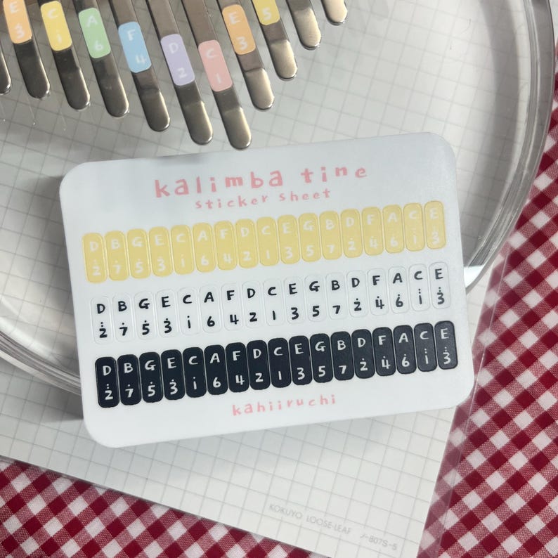 Pu&ograve; includere: Un foglio di adesivi bianco con l'etichetta "kalimba tine" con note musicali e numeri. Il foglio di adesivi ha tre file di note e numeri, con il testo "k&auml;hiiruchi" in basso. Lo sfondo include un kalimba e un motivo a scacchi rossi e bianchi.