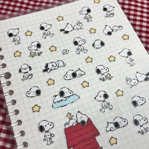 May include: A sheet of stickers featuring Snoopy and Woodstock characters in various poses, along with yellow star shapes. The stickers are on a grid-lined paper, with a red and white checkered background visible.