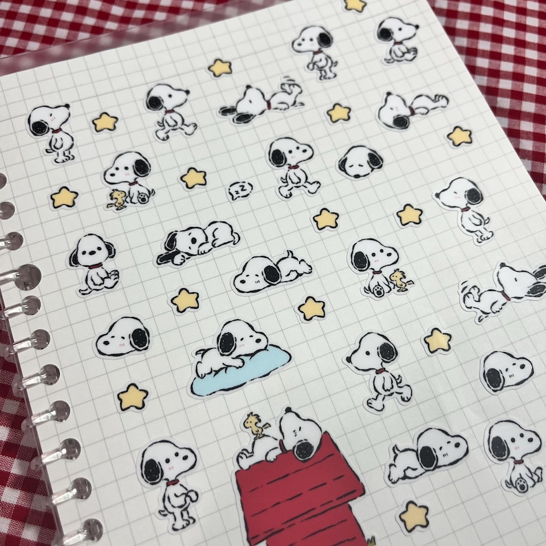 May include: A sheet of stickers featuring Snoopy and Woodstock characters in various poses, along with yellow star shapes. The stickers are on a grid-lined paper, with a red and white checkered background visible.