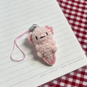 May include: A pink plush axolotl charm with a matching pink lanyard. The axolotl has small black eyes and pink accents on its head and gills. The charm is attached to a silver-colored metal clasp.