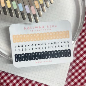 Pu&ograve; includere: Un foglio di adesivi per kalimba, con note musicali e numeri. Il foglio ha il testo "kalimba tine sticker sheet" e il nome "kahiiruchi". Gli adesivi sono per le lamelle del kalimba.