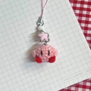 May include: A pink plush charm featuring a cartoon character with red feet and black eyes. The charm is attached to a pink string with a silver clasp, a white bead, and a pink star-shaped charm. The background is a white grid paper.
