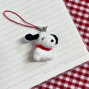 May include: A small, plush white dog charm with black ears and a red collar. The charm is attached to a red cord and silver clasp. The charm is on a lined paper notebook.