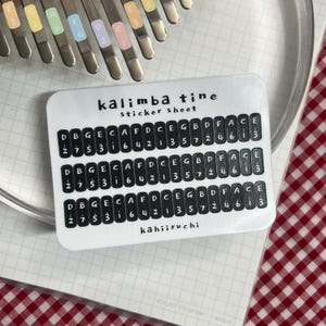 Pu&ograve; includere: Un foglio di adesivi bianco per kalimba, con la scritta "kalimba tine sticker sheet". Il foglio ha file di testo nero con note musicali e numeri. Un kalimba e un panno a scacchi rosso e bianco sono sullo sfondo.