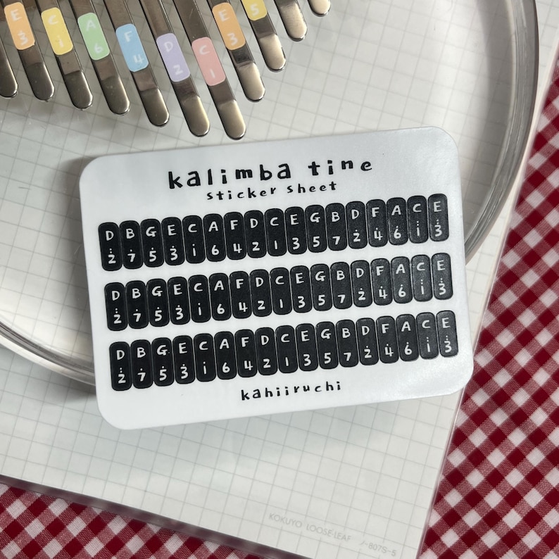 Pu&ograve; includere: Un foglio di adesivi bianco per kalimba, con la scritta "kalimba tine sticker sheet". Il foglio ha file di testo nero con note musicali e numeri. Un kalimba e un panno a scacchi rosso e bianco sono sullo sfondo.
