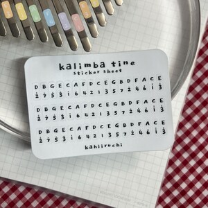 Pu&ograve; includere: Un foglio di adesivi bianco per kalimba, etichettato "kalimba tine sticker sheet". Il foglio presenta file di note musicali e numeri. Uno strumento kalimba &egrave; visibile nella parte superiore. Il foglio di adesivi &egrave; su un quaderno.