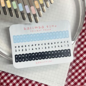 Pu&ograve; includere: Un foglio di adesivi bianco per kalimba, etichettato "kalimba tine sticker sheet". Il foglio presenta file di note musicali e numeri, con il marchio "kahiiruchi" in basso. L'immagine mostra anche le lamelle metalliche di un kalimba.