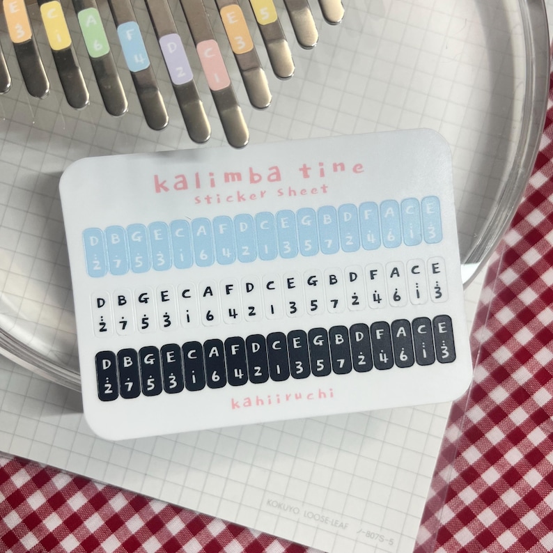 Pu&ograve; includere: Un foglio di adesivi bianco per kalimba, etichettato "kalimba tine sticker sheet". Il foglio presenta file di note musicali e numeri, con il marchio "kahiiruchi" in basso. L'immagine mostra anche le lamelle metalliche di un kalimba.