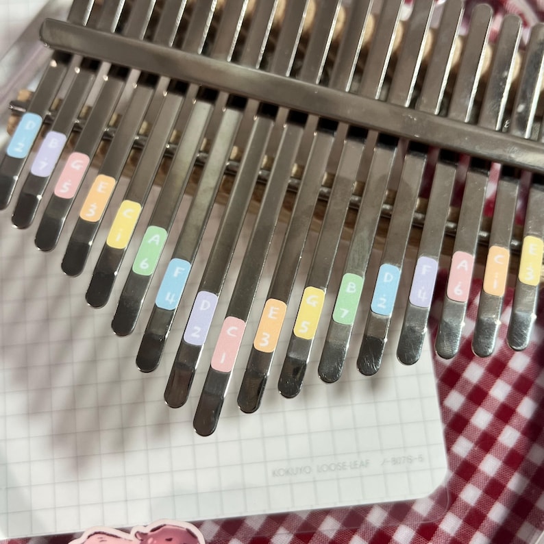 Pu&ograve; includere: Primo piano di un kalimba, uno strumento musicale con lamelle metalliche. Le lamelle sono etichettate con adesivi colorati che indicano le note. Il kalimba poggia su un blocco note con il testo "KOKUYO LOOSE LEAF".