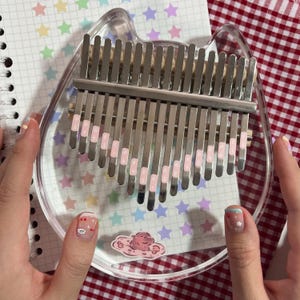 Pu&ograve; includere: Un kalimba trasparente a forma di gatto con lamelle argentate ed etichette rosa. Lo strumento poggia su un quaderno con adesivi a forma di stella. Il kalimba &egrave; tenuto da mani con unghie curate.