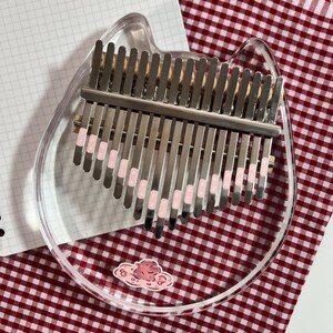 Pu&ograve; includere: Un kalimba in acrilico trasparente a forma di testa di gatto, con lamelle metalliche e dettagli rosa. Lo strumento &egrave; appoggiato su una superficie a scacchi rossi e bianchi, con un adesivo a forma di nuvola.
