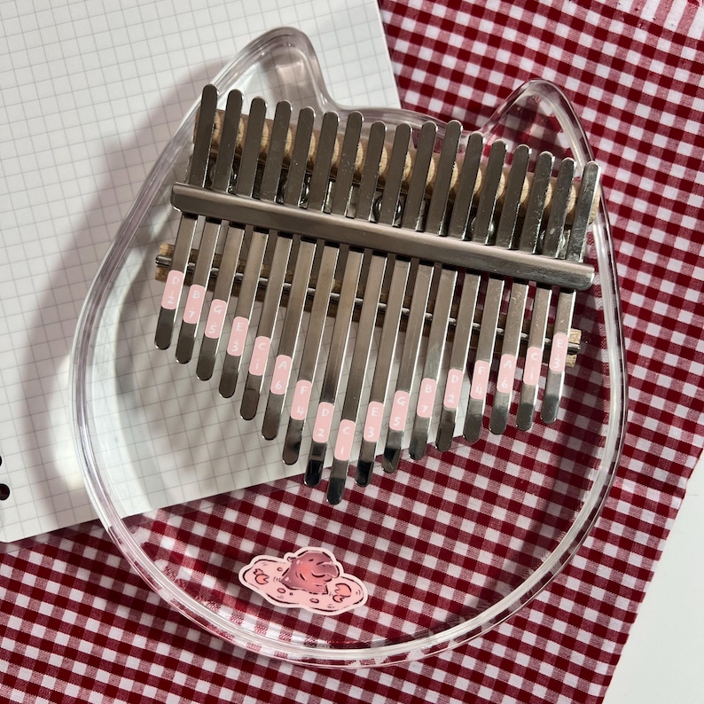 Pu&ograve; includere: Un kalimba in acrilico trasparente a forma di testa di gatto, con lamelle metalliche e dettagli rosa. Lo strumento &egrave; appoggiato su una superficie a scacchi rossi e bianchi, con un adesivo a forma di nuvola.