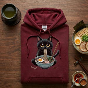 Pode incluir: Moletom bordô com um gato preto de desenho animado comendo ramen em uma tigela azul. O gato tem olhos grandes e amarelos e está segurando pauzinhos. Uma tigela de ramen e uma xícara de chá também são visíveis.