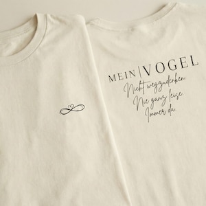 Puede incluir: Dos camisetas color crema. Una camiseta tiene un pequeño símbolo de corazón infinito en el pecho izquierdo. La otra camiseta tiene las palabras "MEIN VOGEL Nicht wegzudenken Nie ganz leise. Immer da" en escritura negra.