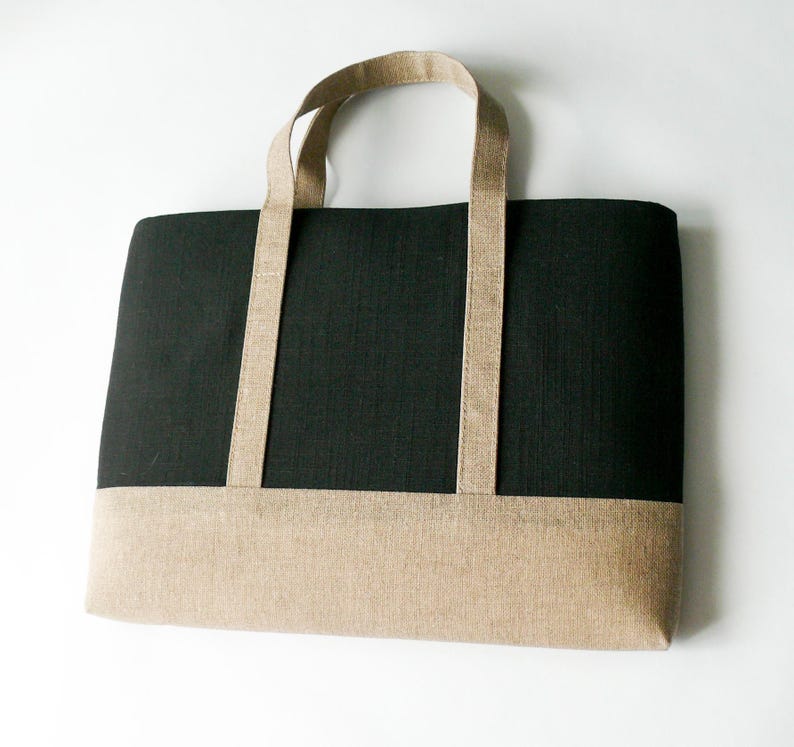 May include: A rectangular tote bag with a black upper section and a tan lower section. The bag has two tan handles and is made of a textured fabric. The bag is designed for carrying items.