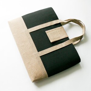 May include: A rectangular tote bag with a geometric design. The bag features a black center panel, a tan side panel, and tan straps. The handle is a loop of the same tan material. The bag is made of a textured fabric.
