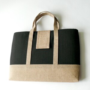 May include: A rectangular tote bag with a black body and a tan bottom panel. The bag has tan handles and a tan flap closure. The bag is made of a textured fabric and is suitable for carrying various items.