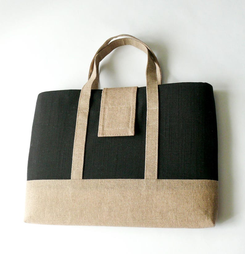 May include: A rectangular tote bag with a black body and a tan bottom panel. The bag has tan handles and a tan flap closure. The bag is made of a textured fabric and is suitable for carrying various items.
