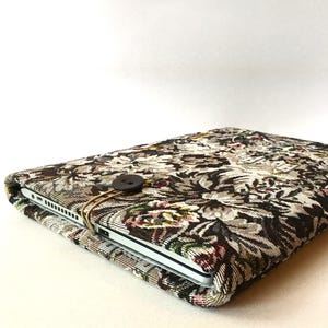 May include: A laptop sleeve made of a brown and beige tapestry fabric with a floral pattern. The sleeve has a brown button closure and a gold metal clasp.