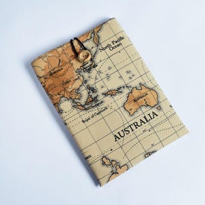 May include: A tan fabric book cover featuring a vintage world map design. The cover is secured with a black elastic loop and a wooden button closure. The map includes continents, oceans, and the word "AUSTRALIA" prominently displayed.