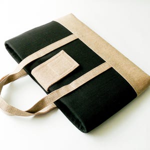 May include: A black tote bag with tan accents. The bag features a rectangular shape with a tan border and straps. A tan rectangular patch is sewn onto the front. The bag has a handle for carrying.