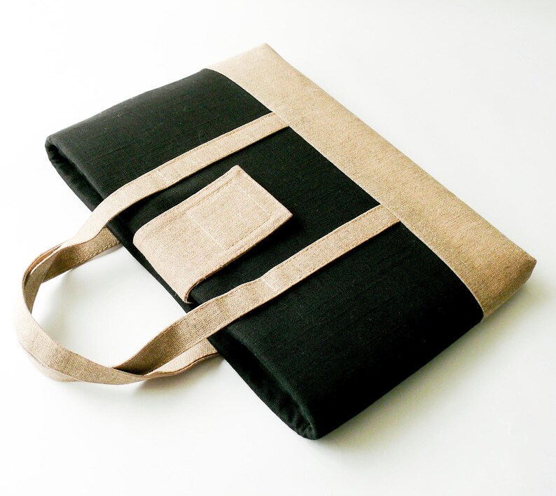 May include: A black tote bag with tan accents. The bag features a rectangular shape with a tan border and straps. A tan rectangular patch is sewn onto the front. The bag has a handle for carrying.