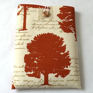 Könnte beinhalten: Cremefarbene Stoffhülle mit einem sich wiederholenden Muster aus rostfarbenen Bäumen und Schrift. Die Hülle wird mit einem Holzknopf und einer weißen Kordelschlaufe verschlossen. Für Tablets oder E-Reader.