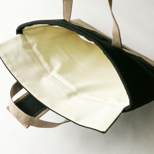 May include: Open view of a tote bag with a black exterior and a beige interior. The bag has two handles and a black trim. The interior lining is a light beige color, and the handles are a light brown.