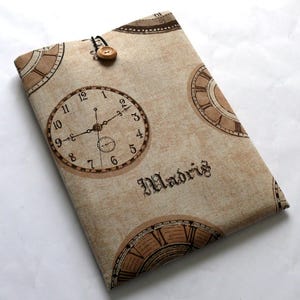May include: A beige fabric laptop sleeve with a repeating pattern of brown clock faces. The sleeve has a brown button closure and the word "Madrid" is printed on the front.