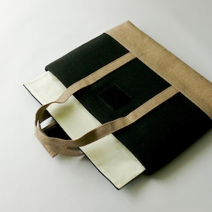 May include: A rectangular tote bag with a black, beige, and white color scheme. The bag features a black central panel, beige trim, and white side panels. The handles are beige, and a small black square is on the front.