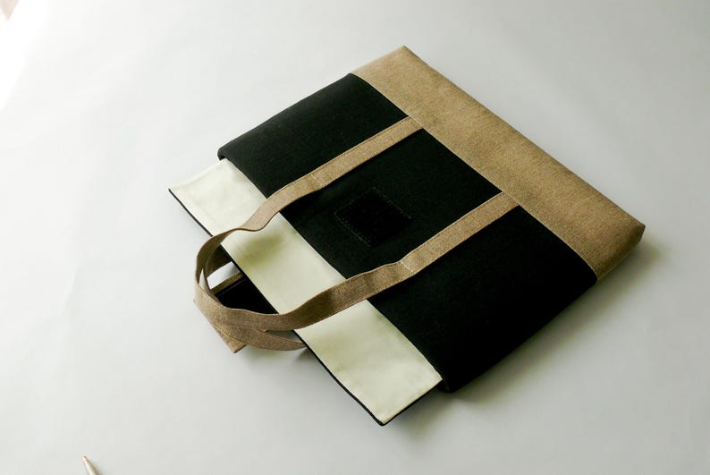 May include: A rectangular tote bag with a black, beige, and white color scheme. The bag features a black central panel, beige trim, and white side panels. The handles are beige, and a small black square is on the front.