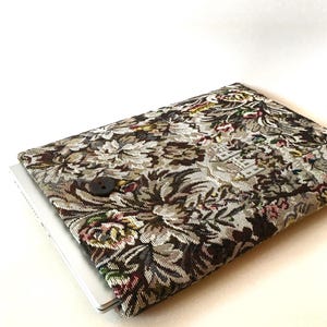 Pode incluir: Uma capa para laptop marrom e bege com um design de tapeçaria floral. A capa tem um fecho de botão único.