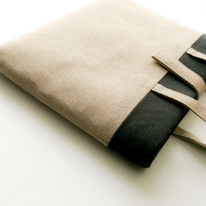 May include: A beige and black tote bag made of a textured fabric. The bag features a rectangular shape with a contrasting black bottom panel and matching beige handles. The bag is designed for carrying items.