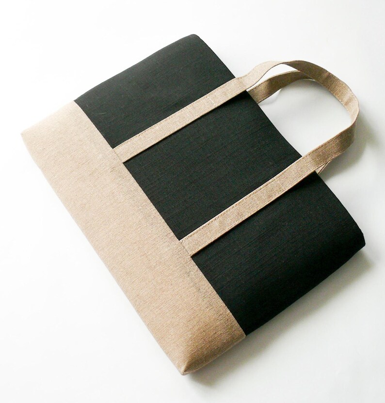 May include: A black and tan tote bag with a rectangular design. The bag features a black center panel and tan-colored top and bottom sections. Tan straps extend from the top of the bag for carrying.