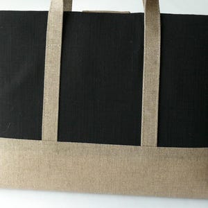 May include: A rectangular tote bag with a black body and a tan bottom panel. The bag has two tan straps and is made of a textured fabric. The bag is designed for carrying items.