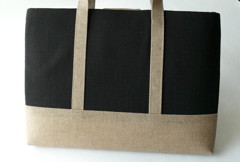 May include: A rectangular tote bag with a black body and a tan bottom panel. The bag has two tan straps and is made of a textured fabric. The bag is designed for carrying items.