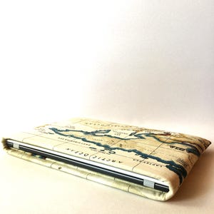 May include: A laptop computer inside a cream-colored case with a vintage world map design. The map features blue ocean details and brown landmasses. The case is closed, showing the top and side.