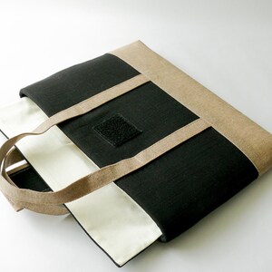 May include: A black and beige tote bag with beige handles and trim. The bag has a square black velcro patch on the front. The interior is white. The bag is made of a textured fabric.