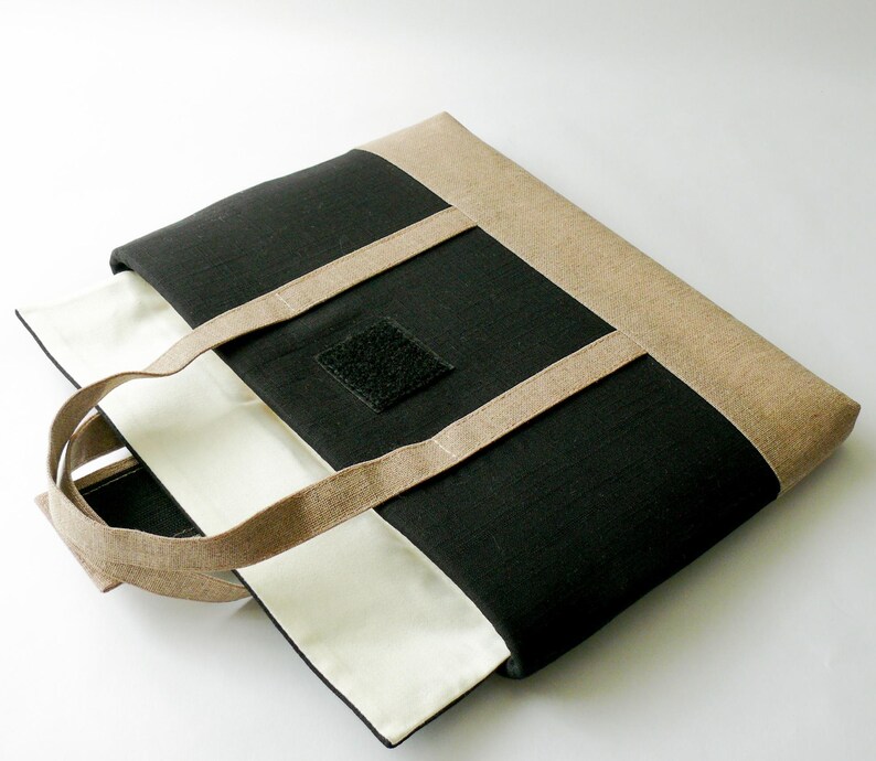 May include: A black and beige tote bag with beige handles and trim. The bag has a square black velcro patch on the front. The interior is white. The bag is made of a textured fabric.