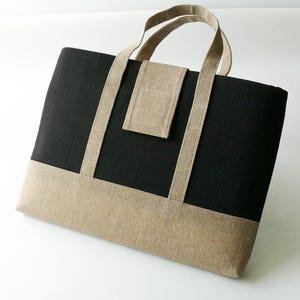 May include: A rectangular tote bag with a black upper section and a tan lower section. The bag has tan handles and a small tan pocket on the front. The bag is made of a textured fabric.