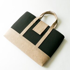 May include: A black and tan tote bag with a rectangular shape. The bag features a tan bottom and accents, with a black center panel. The handles and a small square pocket are also tan. The bag is made of a textured fabric.