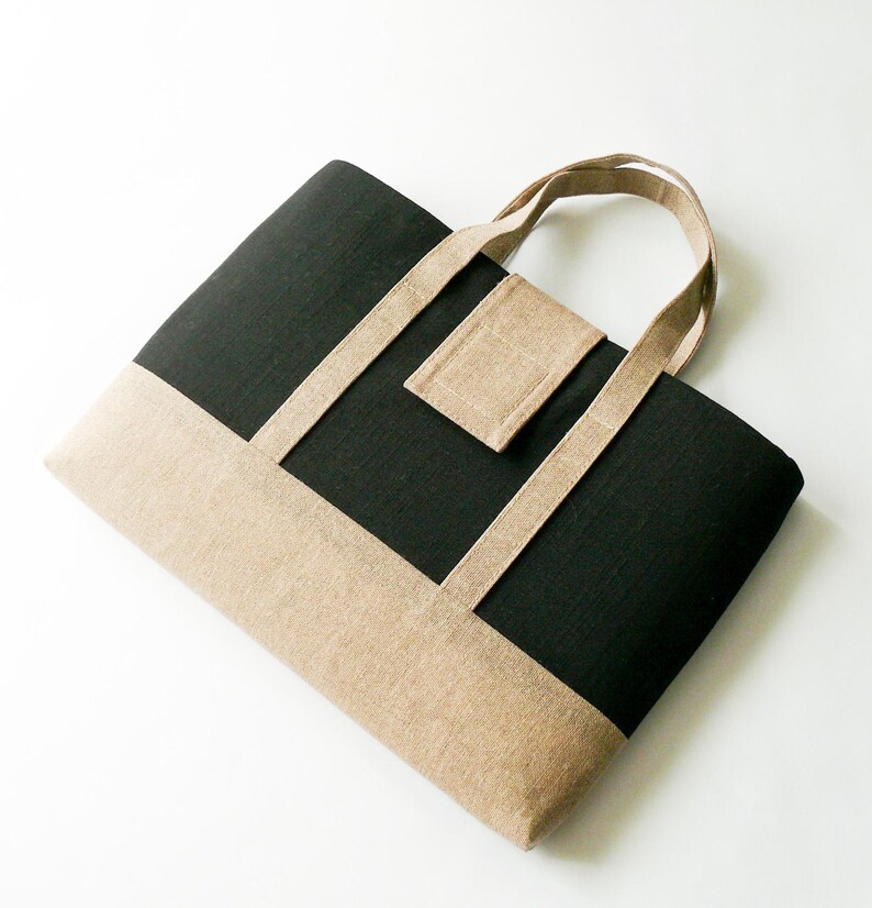 May include: A black and tan tote bag with a rectangular shape. The bag features a tan bottom and accents, with a black center panel. The handles and a small square pocket are also tan. The bag is made of a textured fabric.