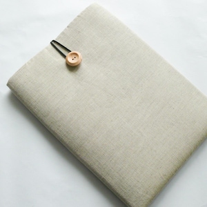May include: A beige linen laptop sleeve with a wooden button fastening. The sleeve is rectangular and has a textured surface. A black elastic loop secures the button. The design is simple and minimalist.