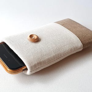 Puede incluir: Una funda para teléfono de lino blanco y marrón con un botón de madera. La funda está diseñada para sostener un teléfono inteligente.