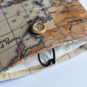 May include: A fabric case with a world map design in beige and brown tones. It features a wooden button and a black elastic loop for closure. The interior is white. The case is designed to hold a small item.