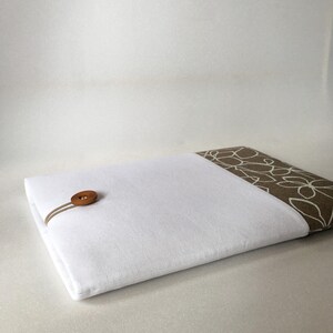 May include: White laptop sleeve with a brown floral pattern on the flap. The sleeve has a brown leather button and loop closure.